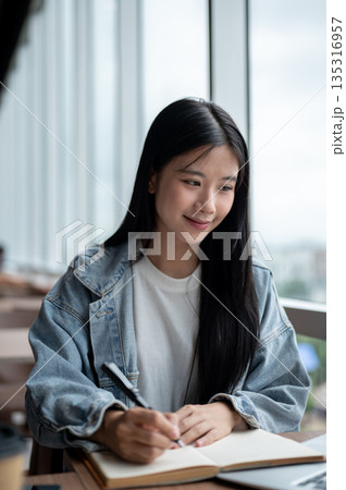 Young asian woman in denim jacket holding fountain pen writing in notebook on wooden table in cafe. Young asian woman in denim jacket holding fountain pen writing in notebook on wooden table in cafe. 135316957