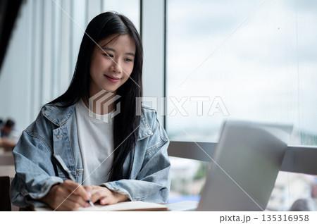Young asian woman in denim jacket holding fountain pen and looking at laptop on wooden table in cafe Young asian woman in denim jacket holding fountain pen and looking at laptop on wooden table in cafe 135316958