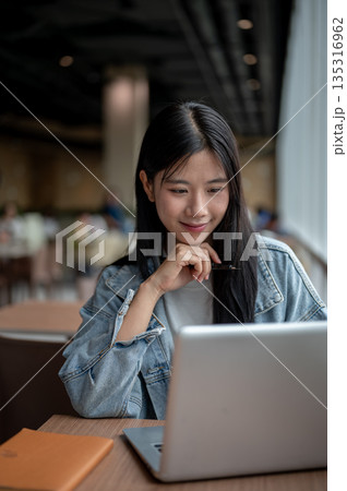 Young asian woman in denim holding pen under chin and smiling as looking at laptop on table in cafe Young asian woman in denim holding pen under chin and smiling as looking at laptop on table in cafe 135316962