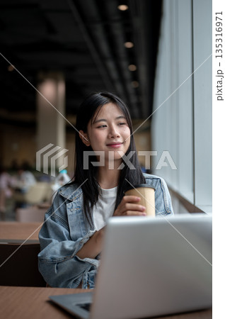 Smiling young asian woman holding coffee while sitting with laptop on table in the cafe or lounge. Smiling young asian woman holding coffee while sitting with laptop on table in the cafe or lounge. 135316971