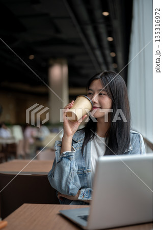 Smiling young asian woman drinking coffee while sitting with laptop on table in the cafe or lounge. Smiling young asian woman drinking coffee while sitting with laptop on table in the cafe or lounge. 135316972