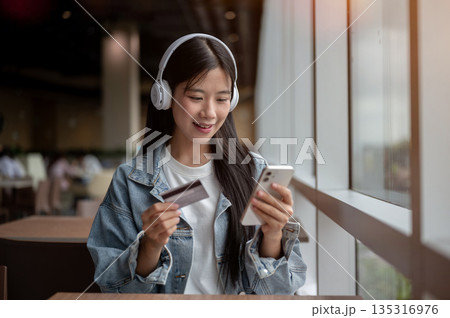 Young asian woman holding credit card and looking at phone while sitting at table in cafe or lounge. 135316976