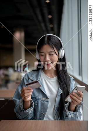 Young asian woman holding phone and looking at a credit card while sitting at table in cafe or lounge. 135316977
