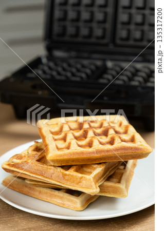 Fresh Pastry on a Plate with a Closed Black Waffle Iron in the Background. Fresh Pastry on a Plate with a Closed Black Waffle Iron in the Background. 135317200