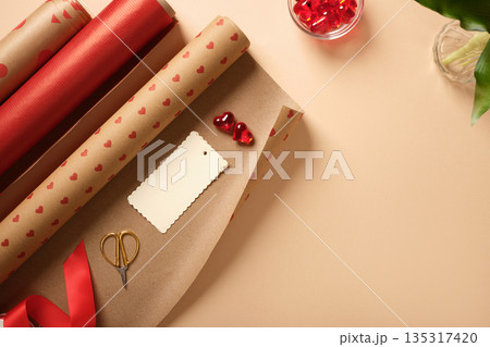 Gift wrapping supplies with red paper rolls, ribbon, scissors, tag and heart decorations on beige background. Copyspace. Gift wrapping supplies with red paper rolls, ribbon, scissors, tag and heart decorations on beige background. Copyspace. 135317420