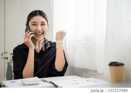 Smiling asian businesswoman or office worker making a fist while talking on phone at working table. 135317490