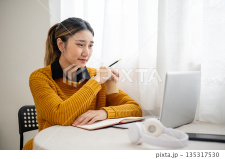 Bored asian woman student office worker holding pencil and coffee while looking at laptop on table. 135317530