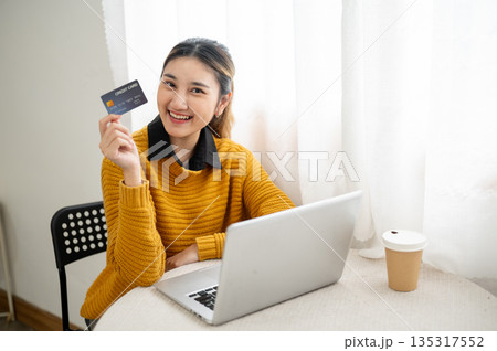 Smiling asian woman holding credit card and showing to camera while sit in front of laptop on table Smiling asian woman holding credit card and showing to camera while sit in front of laptop on table 135317552