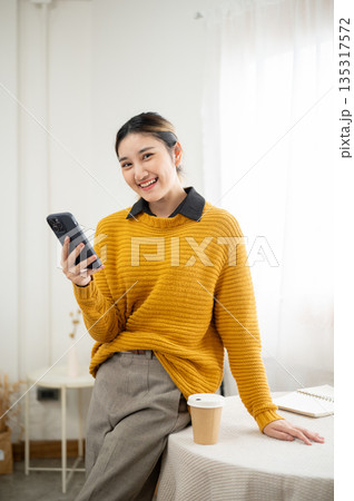 Pretty young asian woman holding and look away from phone while standing leaning against white table Pretty young asian woman holding and look away from phone while standing leaning against white table 135317572