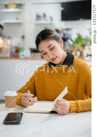 Asian woman holding pencil looking at notebook with phone aside coffee while sitting at cafe's table Asian woman holding pencil looking at notebook with phone aside coffee while sitting at cafe's table 135317577