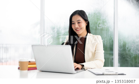 Smiling asian businesswoman or office worker in suit looking and using a laptop at working table. Smiling asian businesswoman or office worker in suit looking and using a laptop at working table. 135318007