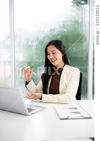 Asian businesswoman or office worker in suit holding pen explaining to a laptop at working table. Asian businesswoman or office worker in suit holding pen explaining to a laptop at working table. 135318012