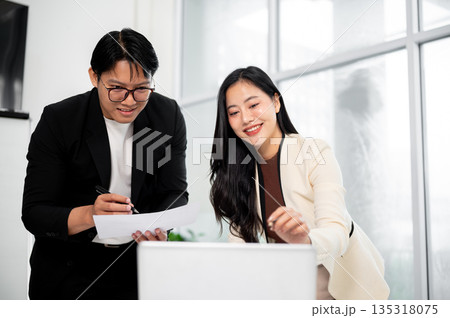 Businesswoman or office worker smiling while pointing on laptop to a man holding document over table Businesswoman or office worker smiling while pointing on laptop to a man holding document over table 135318075