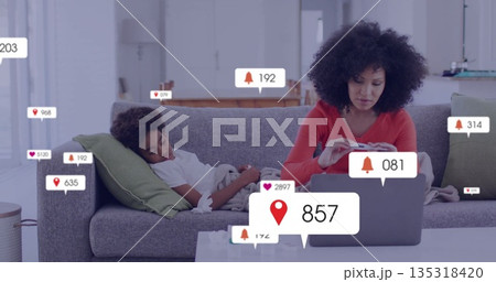 Working mother typing on laptop on sofa in living room, with smartphone, controller and alert icons 135318420