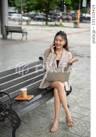 Asian woman talking on phone with hand gesture and sitting cross legged with laptop on bench in park 135318424