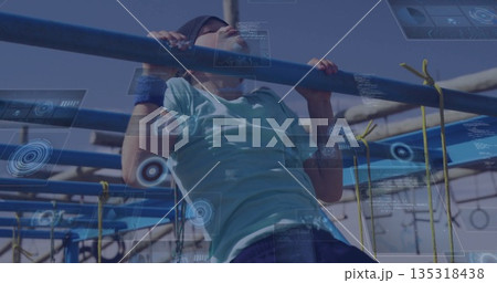 Pulling up boy wearing headband and wristbands on metal bars at obstacle course, yellow rope nets 135318438