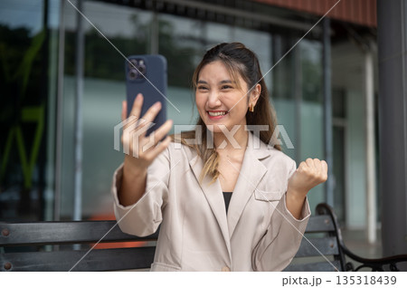 Asian woman holding phone and making a fist looking excited while sitting on bench in front of store Asian woman holding phone and making a fist looking excited while sitting on bench in front of store 135318439