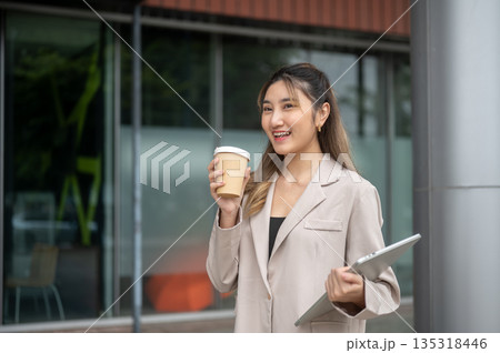 Beautiful asian woman holding coffee cup and carrying a laptop while standing in front of the shop. Beautiful asian woman holding coffee cup and carrying a laptop while standing in front of the shop. 135318446