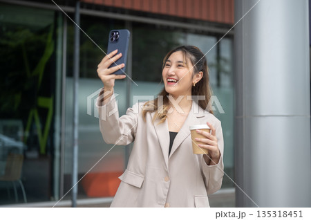 Asian woman holding coffee and taking a photo or video call on phone while standing in front of cafe Asian woman holding coffee and taking a photo or video call on phone while standing in front of cafe 135318451