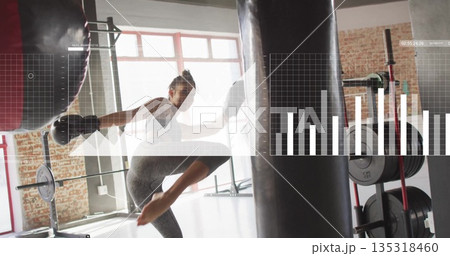 Woman in gray leggings, black gloves kicking and punching heavy bag in gym, with barbell rack 135318460