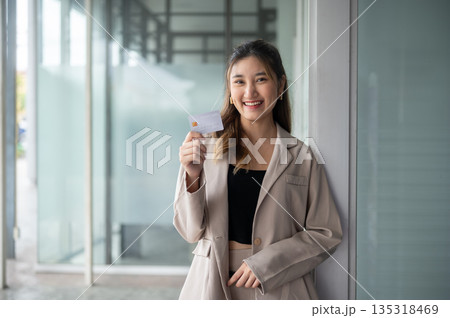 Smiling asian woman standing and holding or showing a credit card in front of a store's glass wall. 135318469