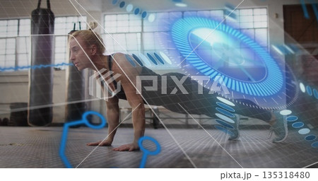 Performing push-up mid-adult woman in athletic wear at gym, with punching bags and HUD graphics 135318480