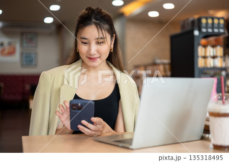 Smiling asian woman holding and looking at phone aside laptop while sitting at wooden table in cafe. 135318495