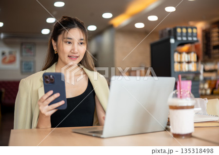 Smiling asian woman holding phone and looking at laptop while sitting at wooden table in the cafe. Smiling asian woman holding phone and looking at laptop while sitting at wooden table in the cafe. 135318498