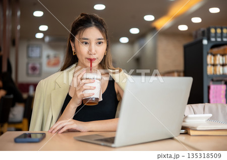 Asian woman enjoy drinking iced tea or coffee while looking at laptop on wooden table in the cafe. Asian woman enjoy drinking iced tea or coffee while looking at laptop on wooden table in the cafe. 135318509