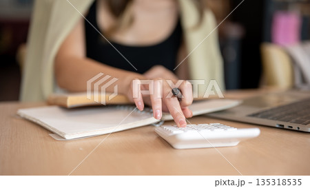 Close up woman's hand holding pen while using calculator aside notebook and laptop on wooden table. Close up woman's hand holding pen while using calculator aside notebook and laptop on wooden table. 135318535