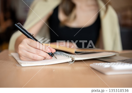 Close up of woman's hand holding pen writing in notebook aside calculator and laptop on wooden table Close up of woman's hand holding pen writing in notebook aside calculator and laptop on wooden table 135318536