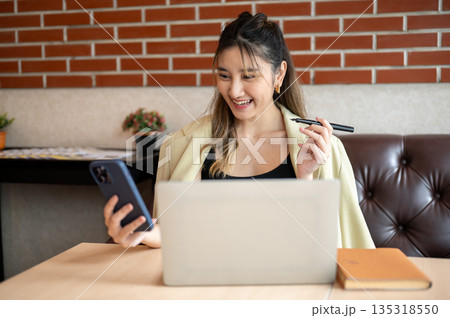 Asian woman holding pen and looking at phone in front of a laptop on table while sit on sofa in cafe Asian woman holding pen and looking at phone in front of a laptop on table while sit on sofa in cafe 135318550