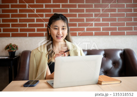 Asian woman thinking as she holding pen pointing at laptop on table while sitting on sofa in a cafe. 135318552