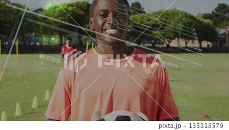 Smiling goalkeeper wearing coral jersey and gloves holding soccer ball on school field, with cones Smiling goalkeeper wearing coral jersey and gloves holding soccer ball on school field, with cones 135318579