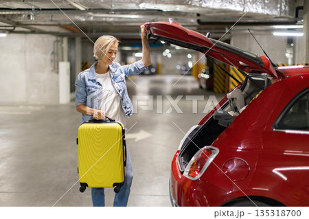 A woman prepares to load her yellow suitcase into the trunk of her red car in a parking garage 135318700