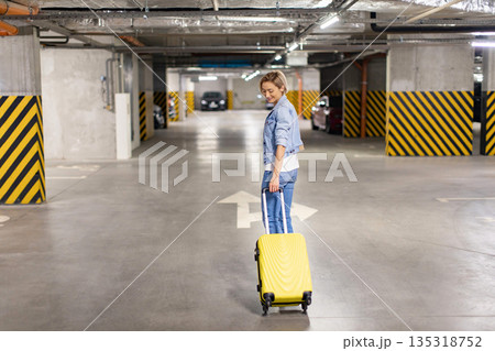 A woman smiles as she pulls a yellow suitcase through an underground parking garage A woman smiles as she pulls a yellow suitcase through an underground parking garage 135318752