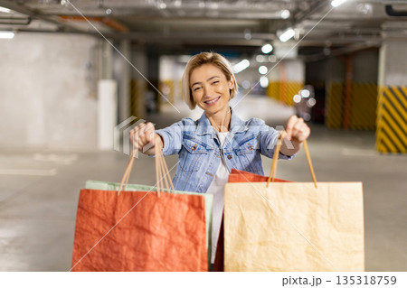 A smiling woman holds shopping bags in a parking garage after a successful shopping trip 135318759