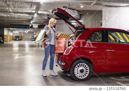 A woman smiles while putting shopping bags into the trunk of her red car in a parking garage 135318760