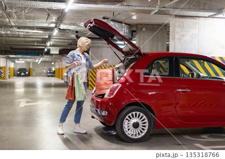 A woman loads shopping bags into the trunk of a red car in an underground parking garage 135318766