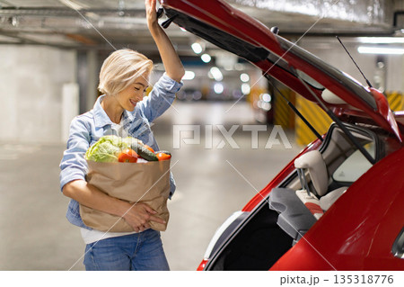 A woman smiles while loading a bag of fresh groceries into the trunk of her car in a parking garage 135318776