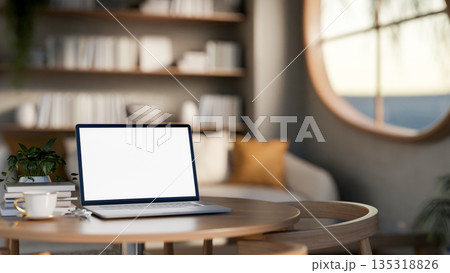 White screen laptop and tea cup on wooden table with armchairs and bookshelf in a sunlit living room 135318826