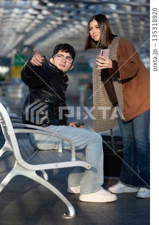 Young man sitting on a bench pointing finger while a woman stands next to him holding a smartphone, both appearing to navigate or seek information at a transit station Young man sitting on a bench pointing finger while a woman stands next to him holding a smartphone, both appearing to navigate or seek information at a transit station 135318920