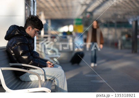 Young man wearing a puffer jacket and glasses is busy on his smartphone while waiting on a bench at a modern train station, a blurred traveler with luggage walking in the background Young man wearing a puffer jacket and glasses is busy on his smartphone while waiting on a bench at a modern train station, a blurred traveler with luggage walking in the background 135318921