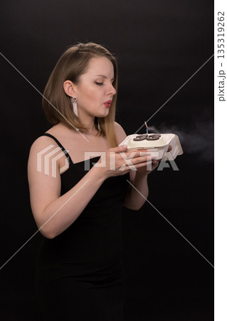 A woman in a black dress blows out a candle on a bento cake. Smoke on a black background. A girl's birthday, happiness, a gift. High quality photo 135319262