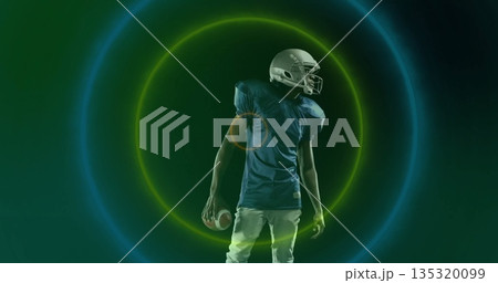 Football player holding football, wearing helmet, jersey with pants in studio with neon rings Football player holding football, wearing helmet, jersey with pants in studio with neon rings 135320099
