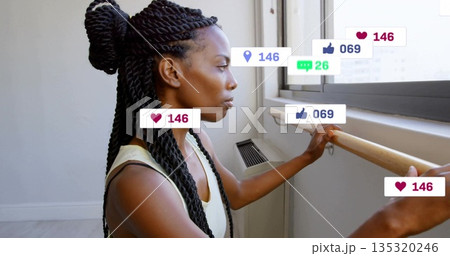 Looking dancer holding barre in tank top by window in studio, with floating reaction icons 135320246