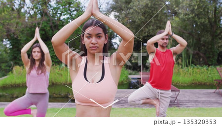 Practicing tree pose woman wearing sports bra guiding yoga on grassy lawn beside pond, with chairs 135320353