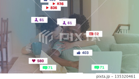 Smiling couple hugging on couch, using laptop browsing with floating icons, holding teal coffee mug 135320461