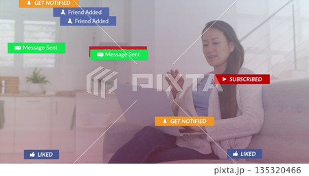 Interacting Korean woman using silver laptop on light grey couch at home, with floating overlays 135320466