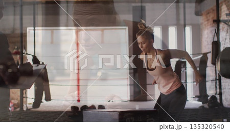 Jumping athlete performing box jump at boxing gym, with plyometric box, ring ropes and heavy bag 135320540